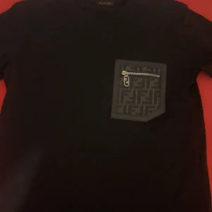 Svart Fendi t-shirt med ficka och dragkedja - Snygg Svart t-shirt från Fendi i bomull med korta ärmar. På bröstet finns en unik ficka med Fendi-logga och dragkedja i silver. Perfekt för dig som gillar exklusiva detaljer och streetstyle. Kom med bud.!😁. Dm för fler information