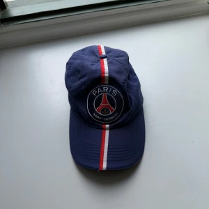 PSG keps med rödvit rand - Mörkblå PSG keps med broderad Paris Saint-Germain-logga framtill och en rödvit rand som går från skärmen över hela kepsen. Justerbar metallspänne bak med PSG-gravyr. Perfekt för dig som älskar fotboll och vill visa ditt lag.