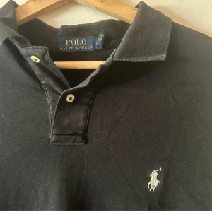 Svart pikétröja från Polo Ralph Lauren - Klassisk svart pikétröja från Polo Ralph Lauren med vit broderad logga på bröstet. Tröjan har korta ärmar, krage och två knappar . Perfekt för en clean och stilren look. Materialet är 100 procent bomull