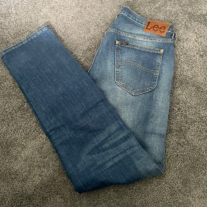 Blå Lee jeans med raka ben - Säljer ett par klassiska blå jeans från Lee med snygg slitning och raka ben. Jeansen har fem fickor, gylf med dragkedja och Lee-lapp bak i midjan. Hör av er vid frågor