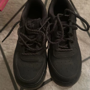 Svarta Nike Air Force 1 sneakers - Klassiska svarta Nike Air Force 1 sneakers med lågt skaft och matchande svarta snören med reflekterande detaljer. Skorna har en robust platt sula, perforerad tå och ikonisk Nike Swoosh i grått på sidorna. Perfekta för dig som gillar streetstyle och tidlös design.