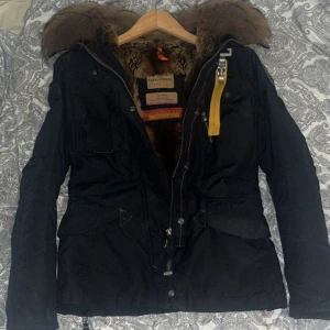 Svart Parajumpers jacka med päls - Svart Parajumpers jacka med stor brun pälskrage och gul detalj vid dragkedjan. Fodrad insida, flera fickor och robust dragkedja framtill. Perfekt för kalla dagar och riktigt snygg streetstil.  .