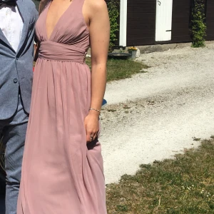 Gammelrosa långklänning med öppen rygg - Säljer en elegant ljuslila långklänning med djup v-ringning och korsade axelband i ryggen. Klänningen har markerad midja och ett svepande, flowy fall. Perfekt för bal eller student! Materialet känns lätt och svalt. En tråd som syns men inget som stör. 