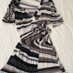 Randig omlottklänning från H&M - Kort omlottklänning från H&M med svart, vit och beige randigt mönster. Klänningen har korta ärmar, v-ringning och knyt i midjan för en snygg passform. Tillverkad i mjukt och lätt material, perfekt för varmare dagar.