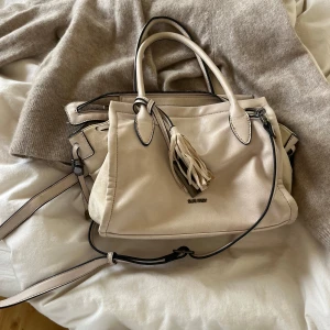 Beige handväska från Suri Frey - Snygg beige handväska från Suri Frey med tofsdetalj framtill. Väskan har både handtag och axelrem, dragkedja upptill och ett rymligt foder med tryckt logga. Perfekt storlek för vardagens essentials och stilren look.