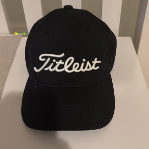 Svart Titleist keps med brodyr - Snygg svart keps från Titleist med vit broderad logga framtill. Justerbar rem baktill för perfekt passform. Klassisk böjd skärm och ventilerande paneler. Perfekt för golf eller streetstyle. Junior keps