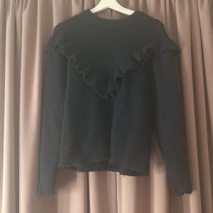 Mörkblå stickad tröja med volang - Mysig mörkblå stickad tröja med lång ärm och snygg volangdetalj som går diagonalt över bröstet och axlarna. Tröjan har en lätt struktur i materialet och en klassisk rund halsringning. Perfekt för dig som gillar en stilren men ändå trendig look.