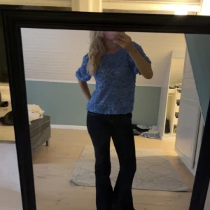 Blå blus med 3D-blommor - Supersöt blå blus med halvtransparent tyg och 3D-blommor över hela toppen. Modellen har rund halsringning och korta puffärmar. Perfekt för dig som vill sticka ut med en unik och lekfull look.