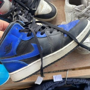 Nike Air Jordan 1 svart/blå sneakers - Säljer ett par Nike Air Jordan 1 sneakers i svart och blått skinn med klassisk Jordan-logga på sidan. Skorna har svarta snören, vit mellansula och svart yttersula. Perfekt för dig som vill sticka ut med en ikonisk och sportig look.