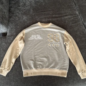 Beige och grå sweatshirt ASOS Design XS - Snygg sweatshirt från ASOS Design i beige och grått med tryck på bröstet och ryggen. Tröjan har långa ärmar, ribbade muddar och coola detaljer som siffran 98 och städer på framsidan samt 'New York' på ryggen. Tillverkad i bomull och polyester.