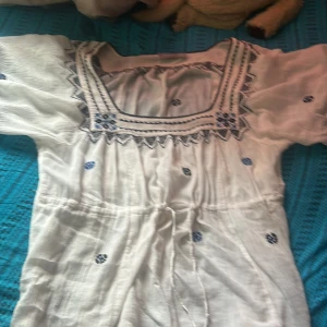 Vit broderad sommarklänning Lindex S - Superfin vit blus från Lindex med blå broderade blommor och snygga detaljer vid halsringningen. Blusen har korta ärmar, knyt i midjan och är gjord i ett lätt bomullstyg – perfekt för varma dagar.