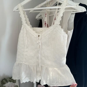 Vit broderad blus med volang - Superfin vit blus med broderade blommor, spetsdetaljer och volangkant nertill. Knappar framtill och breda axelband med spets.
