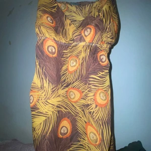 Färgglad omlottklänning med fjädermönster - Säljer en kort omlottklänning mer som linne  från H&M med tropiskt fjädermönster i brunt, gult och orange. Klänningen har v-ringning och resår i midjan för en bekväm passform. Perfekt för dig som gillar färg och mönster!