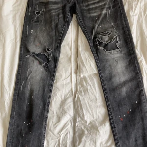 Svarta Dsquared2 jeans med slitningar - Riktigt snygga jeans från Dsquared2, i klassisk svart tvätt med coola slitningar och färgstänk – ett perfekt val för dig som gillar en edgy och trendig look. Jeansen har den karakteristiska Dsquared2-designen med detaljerade slitningar, lappar och råa kanter.  🖤 Färg: Svart tvätt med slitningar och målarstänk 🧵 Design: Distressed med trasiga detaljer och unika inslag 👖 Passform: Slim fit  📏 Storlek: M32/L32 📦 Skick: Använda men i bra skick – inga skador utöver designen 