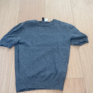 Grå stickad tröja från H&M Divided - En enkel och stilren grå stickad tröja från H&M Divided. Tröjan har korta ärmar, rund halsringning och ribbade muddar vid ärmslut och nederkant. Perfekt att matcha med jeans eller kjol för en clean look.(ny utan prislapp)