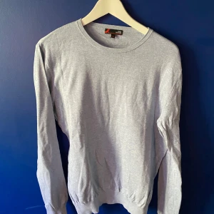Ljusgrå tröja i pima bomull Dressmann - Säljer en stilren ljusgrå tröja från Dressmann i mjuk pima bomull. Tröjan har rund halsringning, långa ärmar och ribbade muddar vid ärmslut och nederkant. Perfekt för dig som gillar en clean och enkel look.