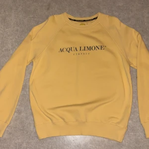 Sweatshirt acqua limone - Snygg gul sweatshirt från Acqua Limone med klassisk logga framtill och stor citronskiva på ryggen. Tröjan har rund hals, långa ärmar och är gjord i mjuk bomull. Perfekt för dig som gillar en clean och fräsch stil. Skicket är väldigt bra då den är använd 1-2 gånger, jag säljer denna för att den är för stor för mig. Inköpspris låg på 1350kr.