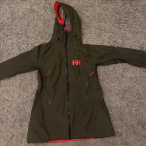 Grön Helly Hansen vindjacka med huva - Snygg grön vindjacka från Helly Hansen med röd HH-logga och detaljer. Jackan har huva, hel dragkedja framtill och två stora fickor med dragkedja. Insidan är grå och materialet är syntet, perfekt för blåsiga dagar. Långärmad och med en clean, sportig look.