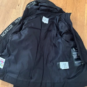 Svart Moncler pufferjacka med huva - Svart pufferjacka från Moncler med huva och tydliga logoband längs insidan. Jackan har dragkedja framtill, Moncler-märke på ärmen och quiltad design. Perfekt för kalla dagar och riktigt snygg streetstil.