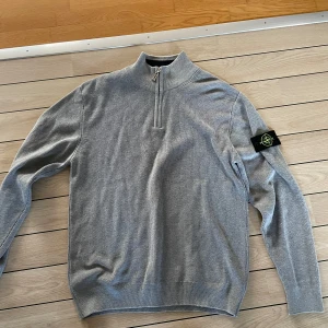 Grå stickad half zip från Stone Island - Snygg grå stickad tröja från Stone Island med klassisk patch på ärmen. Tröjan har half zip i halsen och ribbade muddar. Perfekt för dig som gillar stilren och clean look med en touch av streetwear.