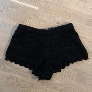 Svarta virkade shorts - Svarta virkade shorts från H&M Divided i storlek XS. De har ett snyggt spetsinspirerat mönster och vågig kant nertill. Shortsen är tillverkade i bomull och har en elastisk midja för skön passform. Perfekta för sommaren och festivaler!