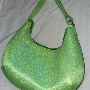 Grön glittrig axelväska - Snygg axelväska i glittrande limegrönt syntetmaterial. Väskan har en rundad form, justerbar axelrem och stängs med dragkedja. Insidan är ljus med en liten innerficka för småsaker. Perfekt för att addera färg till din outfit!