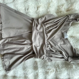 Beige kort omlottklänning med knyt - Supersöt beige topp med omlott och knyt vid axeln. Den har en lätt utsvängd nederdel och markerad midja. Mjuk och skön i bomullsmaterial, perfekt för varma dagar. Stilren och enkel att matcha med accessoarer.