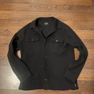 Svart overshirt från Only & Sons - Stilren svart overshirt från Only & Sons med klassisk krage, två bröstfickor med knappar och långa ärmar. Perfekt lager-på-lager-plagg med enkel knäppning framtill. Snygg och enkel att matcha till det mesta.