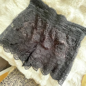 Svarta virkade spets-shorts - Superfina svarta shorts med bred resår i midjan och detaljerad virkad spets över hela plagget. De har ett blommigt mönster i spetsen och vågig kant nertill. Perfekta för sommarens varma dagar och ger en boho-vibe till outfiten.