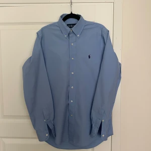 Blå skjorta från Ralph Lauren S - Klassisk ljusblå skjorta från Ralph Lauren i bomull, custom fit. Skjortan har button down-krage, långa ärmar och den ikoniska broderade loggan på bröstet. Perfekt för en clean och stilren look. Sjukt skön skjorta under sommaren! 