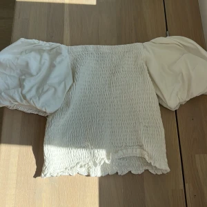 Vit blus med puffärmar från Gina Tricot - Supersöt vit blus från Gina Tricot med smockad kropp och stora puffärmar. Toppen har en croppad passform och volangkant nertill. Perfekt för dig som gillar romantisk och trendig stil.