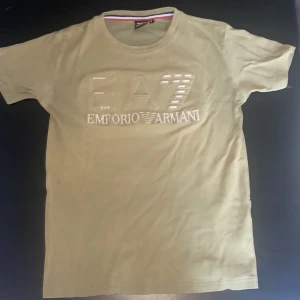 Beige EA7 Emporio Armani t-shirt S - Snygg beige t-shirt från EA7 Emporio Armani med stort logotryck framtill. Klassisk rund halsringning och korta ärmar. Tillverkad i mjuk bomull som känns skön mot huden. Perfekt för dig som gillar stilrena och exklusiva plagg.