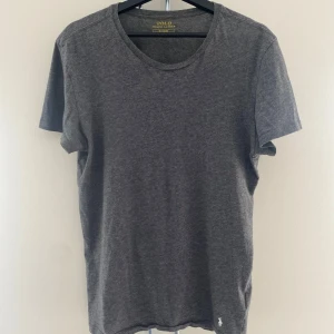 Mörkgrå Ralph Lauren T-shirt - Snygg mörkgrå t-shirt från Polo Ralph Lauren med klassisk passform och rund hals. Tillverkad i mjuk bomull och prydd med den ikoniska vita broderade loggan vid nederkanten. Perfekt basic-plagg med stilren look.