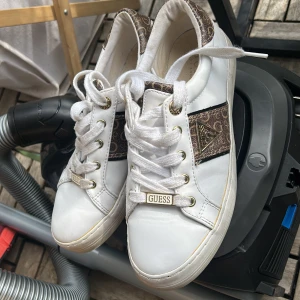 Vita sneakers från Guess med logomönster - Snygga vita sneakers från Guess med bruna detaljer och logomönster på häl och sidor. Skorna har guldfärgade metalldetaljer, klassisk snörning och Guess-logga både fram och bak. Perfekta för dig som gillar en trendig och sportig look.