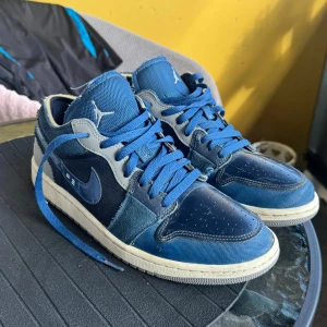 Nike Air Jordan 1 Low OG Blå US9 - Säljer ett par Nike Air Jordan 1 Low OG Limited edition i olika blå nyanser med vita och grå detaljer. Skorna har snörning, vit sula och klassisk Air Jordan-logga på hälen. Materialmix av mocka, skinn och textil ger en cool look. Perfekta för dig som gillar streetwear och sneakers.
