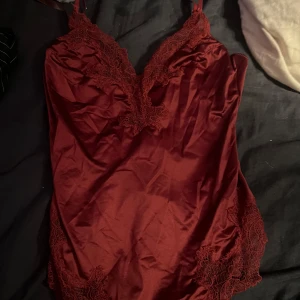 Vinröd satin linnebody med spets - Supersnygg vinröd linnebody i satin med tunna axelband och v-ringning. Dekorerad med matchande spetsdetaljer längs urringning och sidorna för en extra lyxig känsla. Perfekt att styla med jeans eller kjol för en trendig look.