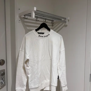 Vit tröja från Palm Angels - Säljer en stilren vit tröja från Palm Angels med deras ikoniska logga i svart och rosa på baksidan. Tröjan har långa ärmar och en klassisk rund halsringning. Perfekt för en avslappnad och trendig look. helt oanvänd