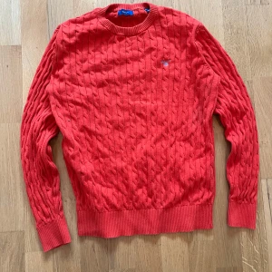 Röd kabelstickad tröja från GANT - Säljer en snygg röd kabelstickad tröja från GANT. Tröjan har en klassisk rund halsringning och ett broderat GANT-emblem på bröstet. Perfekt för en stilren look. storlek 158/164 cm.