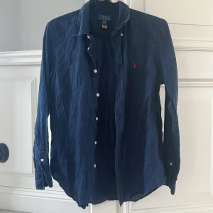 Blå Ralph lauren skjorta - Säljer en klassisk blå skjorta från Polo Ralph Lauren med lång ärm och knappar framtill. Skjortan är 100% linne och är bara använd en säsong vilket gör att den är i topp skick. Storlek: L (14-16). Skriv vid funderingar!!