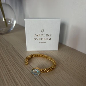 Guldarmband från Caroline Svedbom - Elegant guldarmband från Caroline Svedbom med en stor, glittrande sten i centrum. Armbandet har en flätad design som ger en lyxig känsla. Perfekt för att lägga till en touch av glamour till din outfit.
