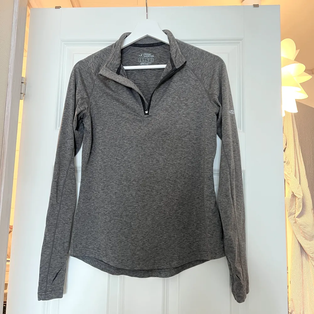 Säljer en grå långärmad tröja med halv dragkedja. Perfekt för träning med sitt DRY-PLUS material som håller dig torr och bekväm. Enkel och stilren design med logga på ärmen.. Sportswear.