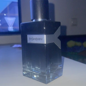 Ysl Y edp  - Elegant parfym från Yves Saint Laurent i en stilren flaska med mörkblått glas och en metallisk etikett. Flaskan har en modern design med en svart kork och tydliga linjer. Perfekt för den som söker en sofistikerad doft.