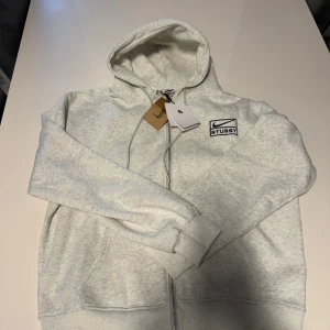 Grå hoodie från Nike x Stussy - Säljer en stilren grå hoodie från Nike i samarbete med Stussy. Tröjan har en dragkedja framtill och en klassisk huva. Perfekt för en avslappnad stil med en touch av streetwear. Passar både för vardag och chill. Säljes pga den var för stor, storlek M fast stor i storleken! 