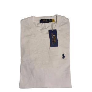 Vit t-shirt från Ralph Lauren - Säljer en klassisk vit t-shirt från Ralph Lauren med den ikoniska loggan broderad i blått på bröstet. Perfekt för en stilren och avslappnad look. T-shirten har en normal passform och är tillverkad i mjuk bomull.