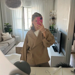 Beige kappa från Dagmar - Säljer en stilren beige kappa från Dagmar, perfekt för höst. Jackan har en unik omlott-design med knappar och ett bälte i midjan. Tillverkad i en ullblandning som ger en lyxig känsla. Passar perfekt för att hålla stilen när det blir kallare.  Knappen på ena armen är sönder. Nypris 6000kr. 