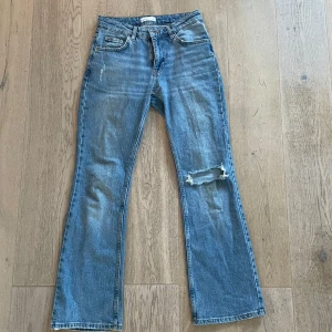 Blå bootcut jeans - Säljer ett par blå bootcut jeans med en snygg slitning på ena benet. De har en klassisk femficksdesign och är perfekta för en avslappnad stil. Högmidjade och i ett mjukt denimtyg.