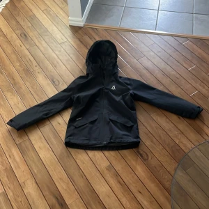Svart vindjacka från Haglöfs - Säljer en svart vindjacka från Haglöfs med huva och dragkedja. Perfekt för utomhusaktiviteter med sin funktionella design. Jackan har långa ärmar och en stilren look.passar folk mellan 145-153