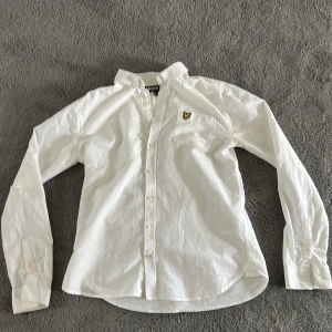 Vit skjorta från Lyle & Scott - Säljer en stilren vit skjorta från Lyle & Scott med klassisk krage och knappar framtill. Skjortan har ett broderat emblem på bröstet och långa ärmar. Perfekt för en snygg och avslappnad look.