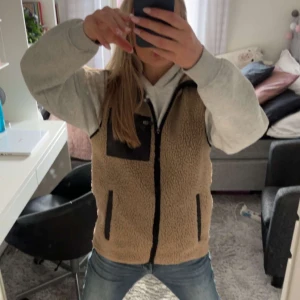 Beige teddyväst med dragkedja - Säljer en mysig beige teddyväst med dragkedja och svarta detaljer. Västen har två praktiska fickor framtill och en bröstficka med dragkedja. Perfekt för lager-på-lager under kyliga dagar. Men även under sena vår/sommarkvällar☀️🤗