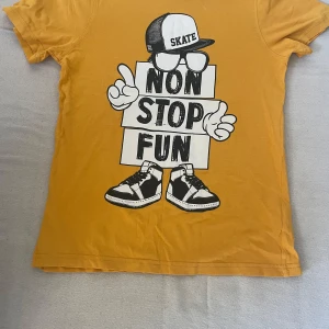 Gul t-shirt med tryck - Cool gul t-shirt med ett stort tryck av en figur med keps och sneakers som håller en skylt med texten 'Non Stop Fun'. Perfekt för en avslappnad stil.